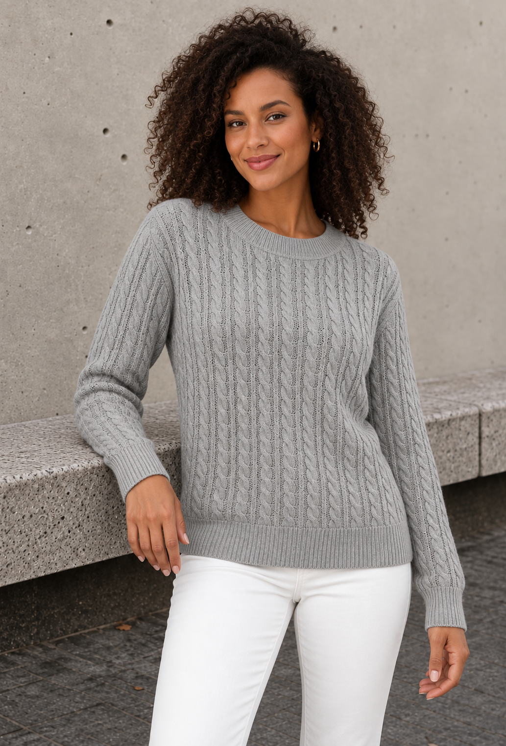 Women's grey cable knit sweater with ribbed crew neck and hem, leaning on a bench